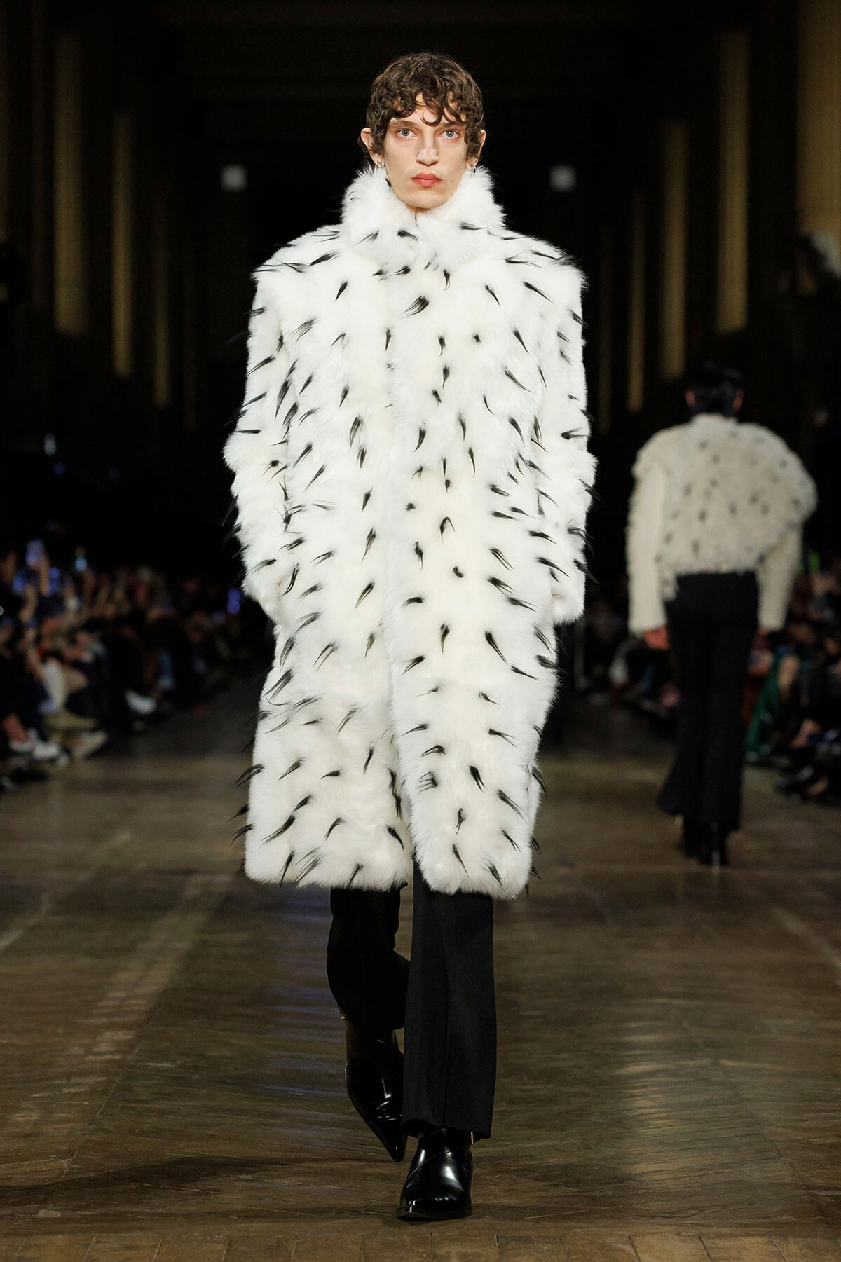 Model walks the runway in a long white fur coat with black feather-like markings, paired with black trousers and glossy boots.