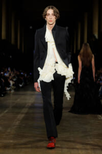 Male model on a runway in a black blazer over a cream ruffled blouse, black trousers, and red patent shoes. Audience visible in background.