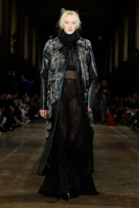 Model walks a runway in a long black embellished coat with silver beadwork over sheer black gown and ruffled collar.