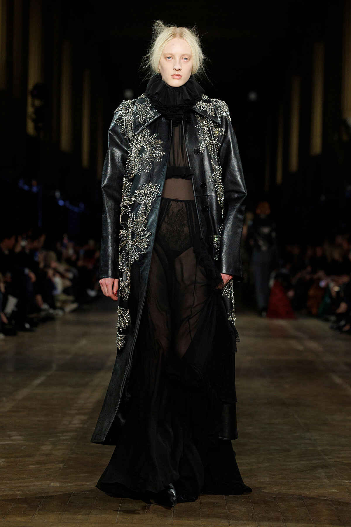 Model walks a runway in a long black embellished coat with silver beadwork over sheer black gown and ruffled collar.