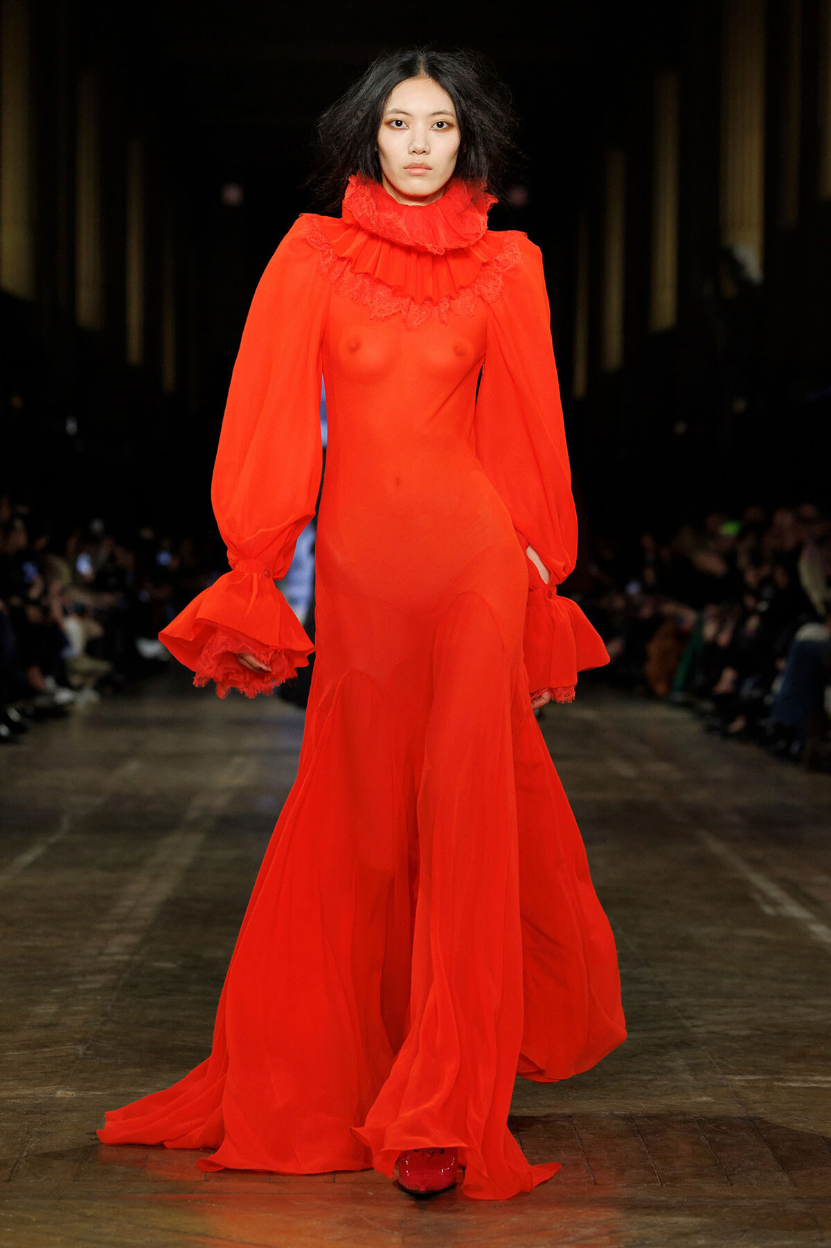Model walks the runway wearing a sheer, bright red gown with a high ruffled collar and voluminous, trumpet sleeves that flare at the wrists, the dress flowing to the floor.