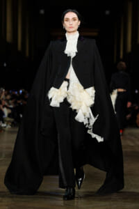Runway model in a black cape over a white ruffled blouse and black trousers.