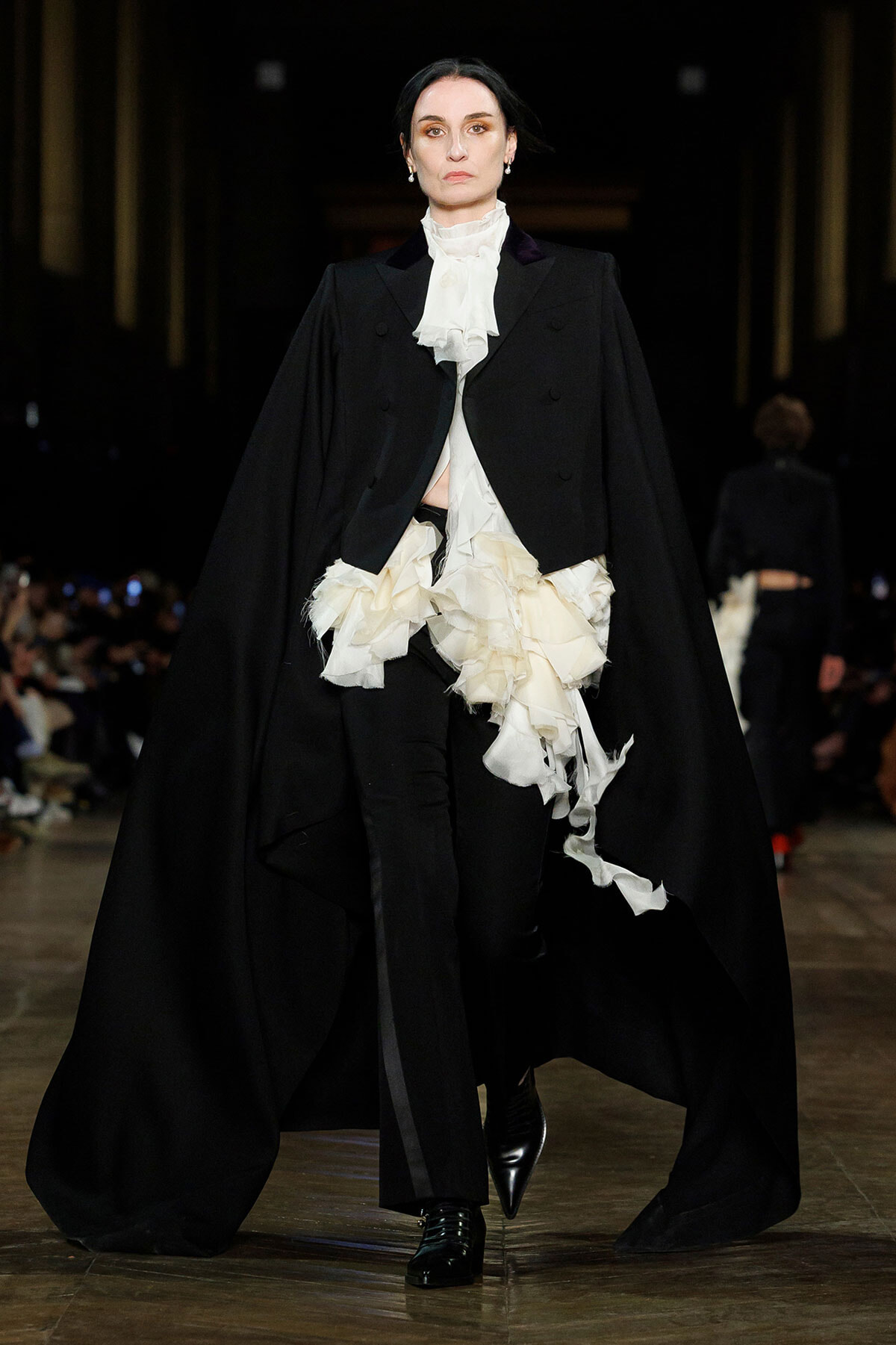 Runway model in a black cape over a white ruffled blouse and black trousers.