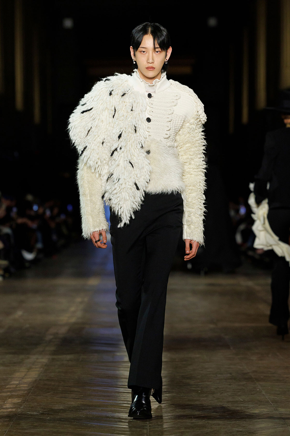 Runway model wears a voluminous white textured coat draped over one shoulder, paired with black trousers and boots.