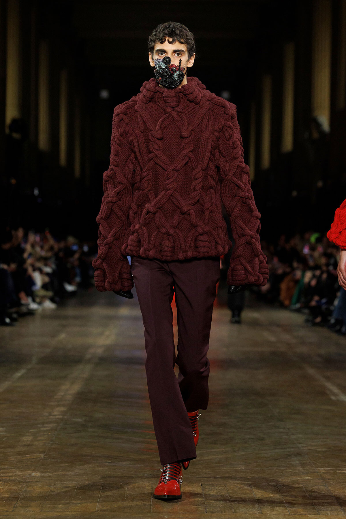 Male model walks a fashion show in a chunky burgundy cable-knit turtleneck with matching maroon pants and red lace-up boots, wearing a floral-pattern mask.