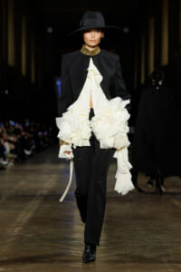 Model on runway wearing a black tailored suit with a dramatic white ruffled blouse, plus a wide-brim hat and gold neckpiece.