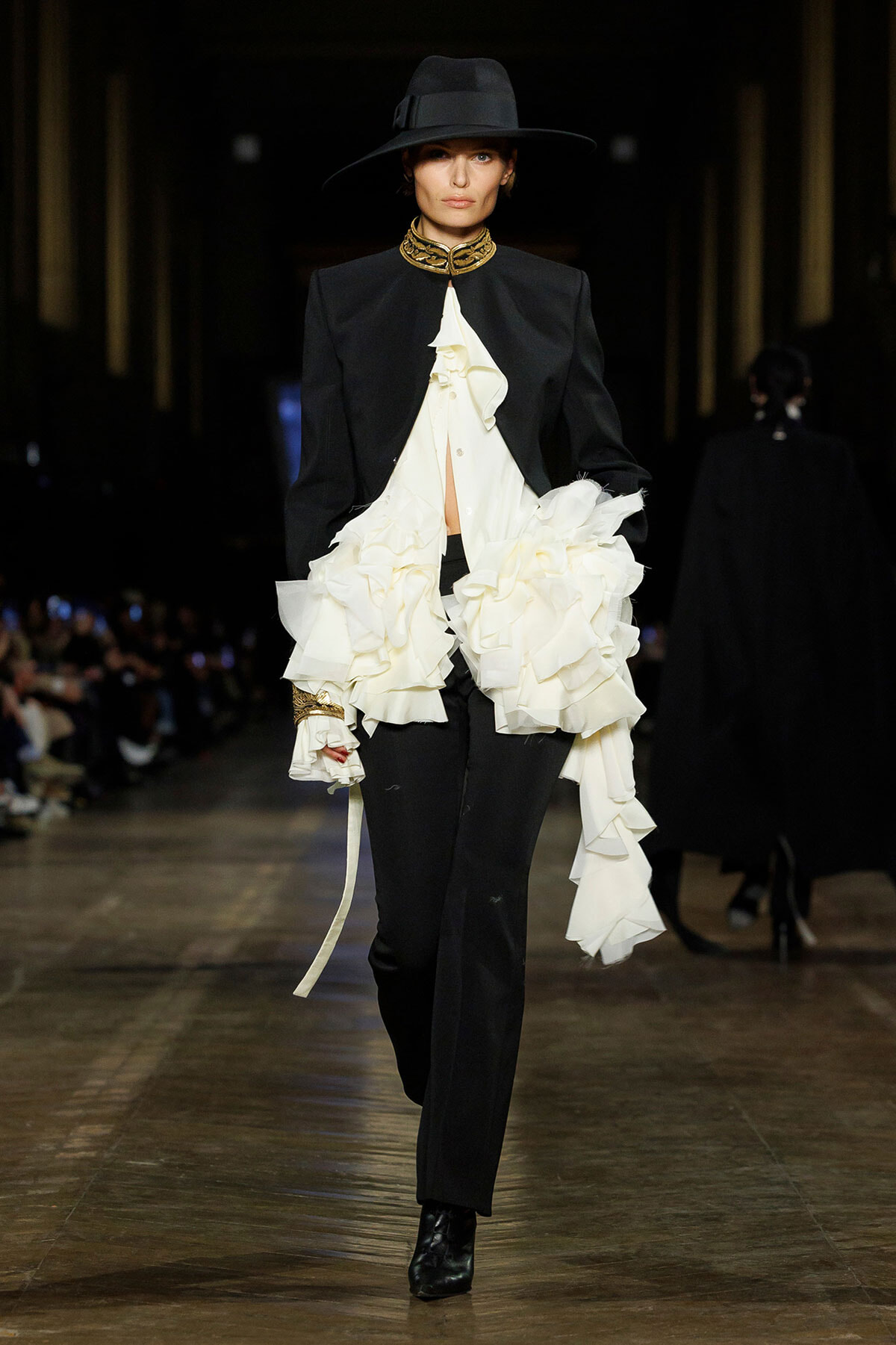 Model on runway wearing a black tailored suit with a dramatic white ruffled blouse, plus a wide-brim hat and gold neckpiece.