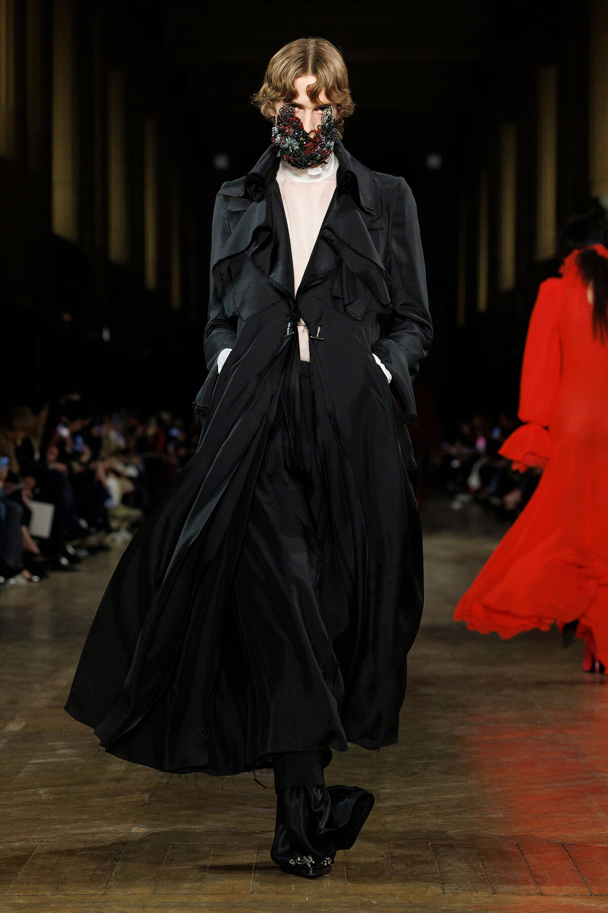 Model on a runway wearing a voluminous black trench with ruffles and a jeweled face mask, hands in pockets.