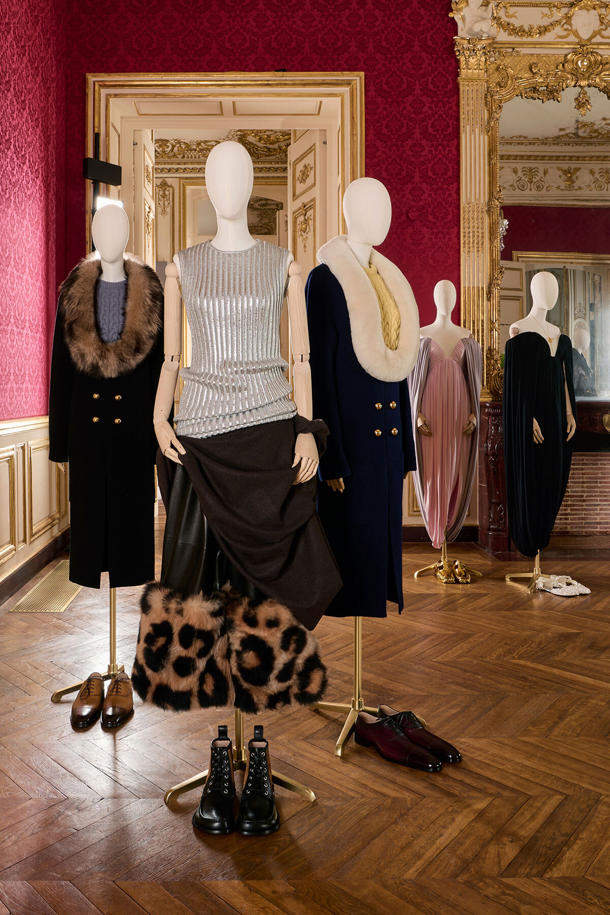 Mannequin display in a luxurious red and gold room showing fur coats, sequin top, and long dresses with boots on the floor nearby.