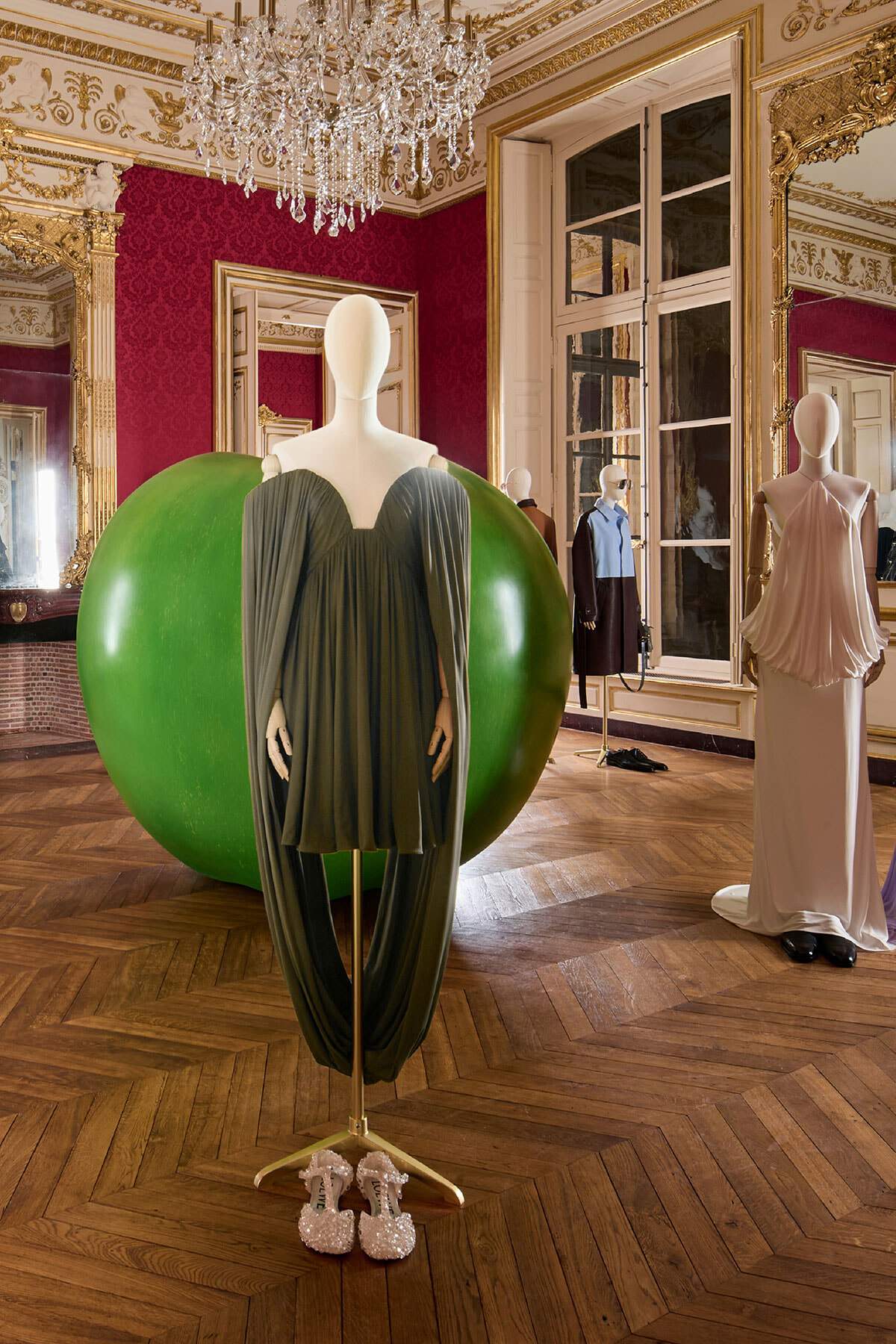 Mannequin in a dark green draped gown in front of a large green sphere in an ornate ballroom with a crystal chandelier above.