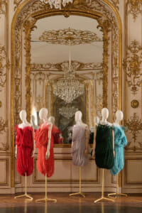 Elegant room with gilded ornate walls and crystal chandeliers, displaying five mannequin-dressed gowns in red, coral, lavender, charcoal, green, and teal hues.