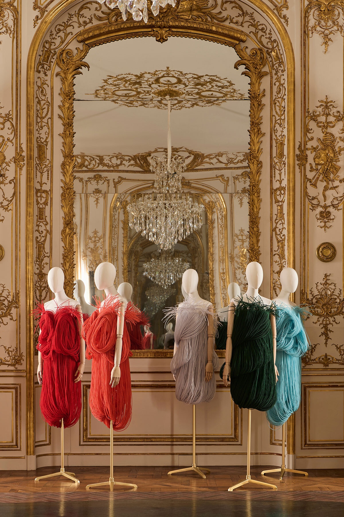 Elegant room with gilded ornate walls and crystal chandeliers, displaying five mannequin-dressed gowns in red, coral, lavender, charcoal, green, and teal hues.