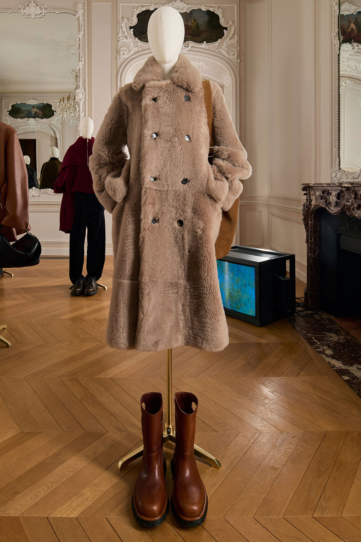 Mannequin in a beige faux‑fur double‑breasted coat, with a brown bag and brown boots beneath on a wooden floor in a showroom.