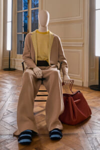 Seated mannequin wearing a beige suit with a yellow striped top, brown wide-leg pants, and a red leather bag nearby.