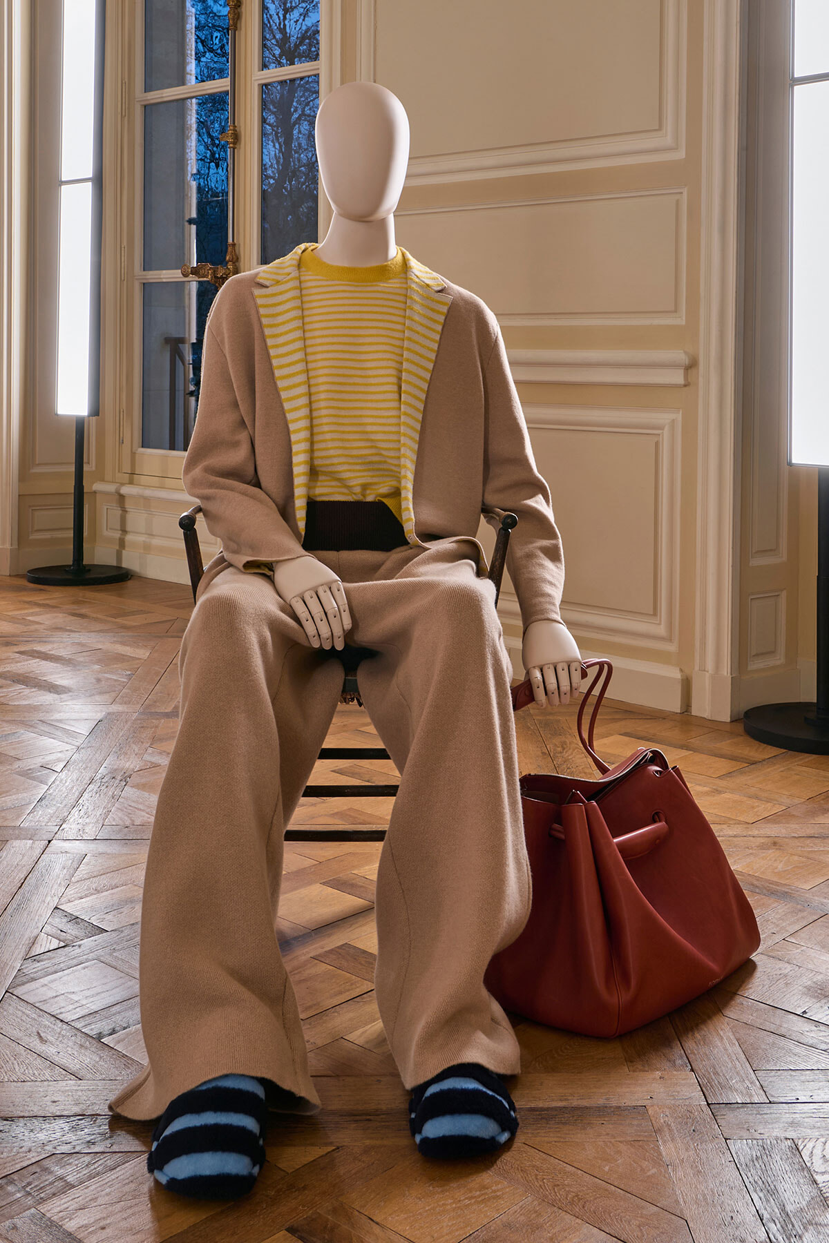 Seated mannequin wearing a beige suit with a yellow striped top, brown wide-leg pants, and a red leather bag nearby.