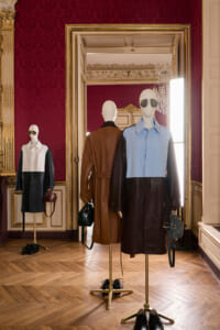 Mannequins wearing color-block coats in a gilded, red-walled showroom with a large ornate mirror behind them.