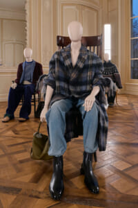 Mannequin in a blue plaid coat, jeans and black boots sits in a decorative room with ornate moldings; olive bag on the floor beside it.