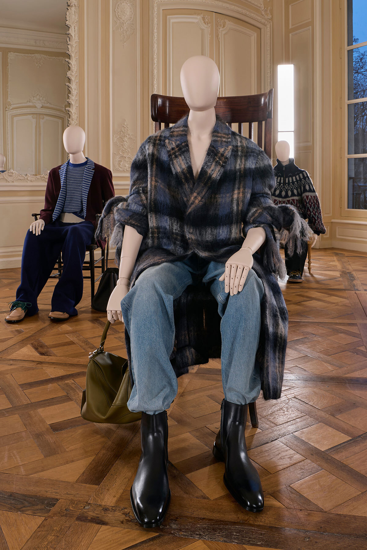 Mannequin in a blue plaid coat, jeans and black boots sits in a decorative room with ornate moldings; olive bag on the floor beside it.