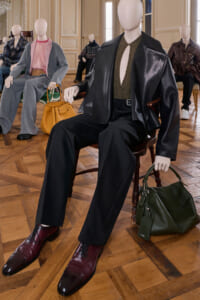 Mannequin wearing a black leather jacket, olive knit top, and wide black pants sits with burgundy shoes nearby and a green handbag on the floor in a fashion showroom.
