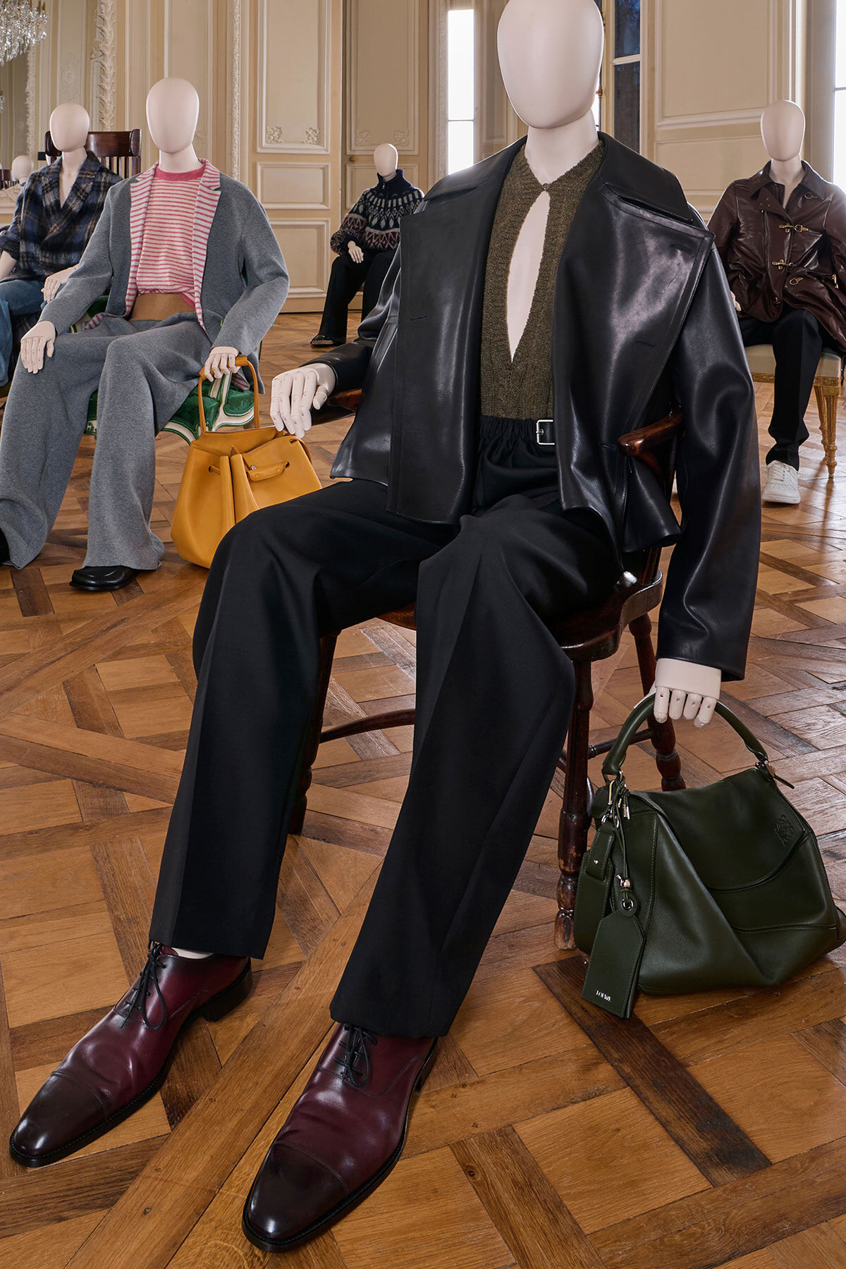 Mannequin wearing a black leather jacket, olive knit top, and wide black pants sits with burgundy shoes nearby and a green handbag on the floor in a fashion showroom.