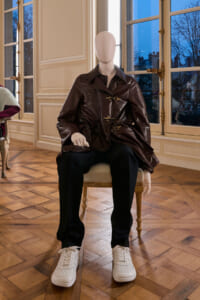 Mannequin wearing a dark brown leather coat, black pants, and white sneakers seated on a gilded chair in a bright, paneled room with large windows.