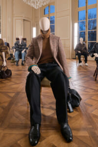Male mannequin seated in a chic room wearing a brown turtleneck, plaid blazer, black trousers, and black shoes with a teal bracelet; a black bag rests beside him.