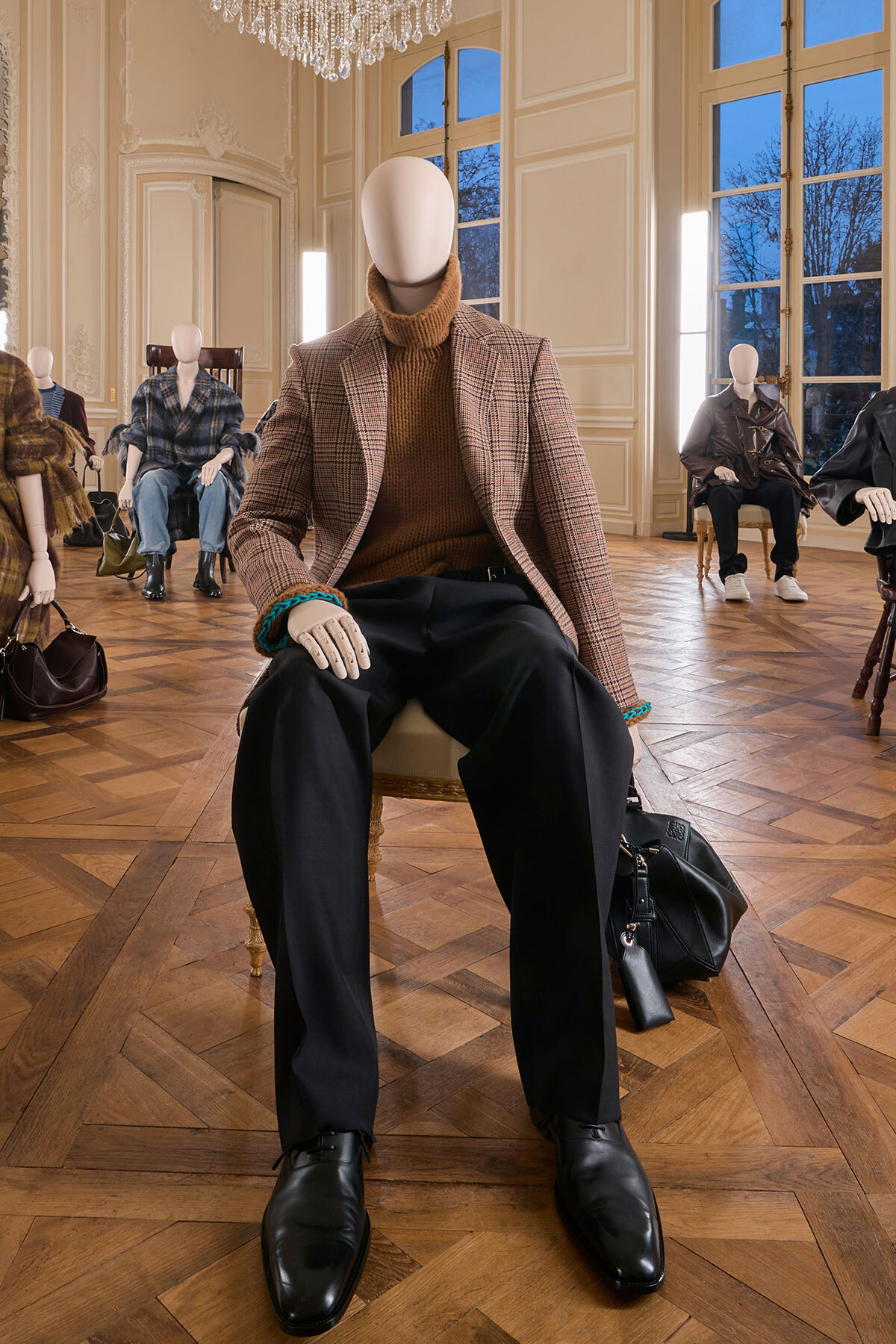 Male mannequin seated in a chic room wearing a brown turtleneck, plaid blazer, black trousers, and black shoes with a teal bracelet; a black bag rests beside him.