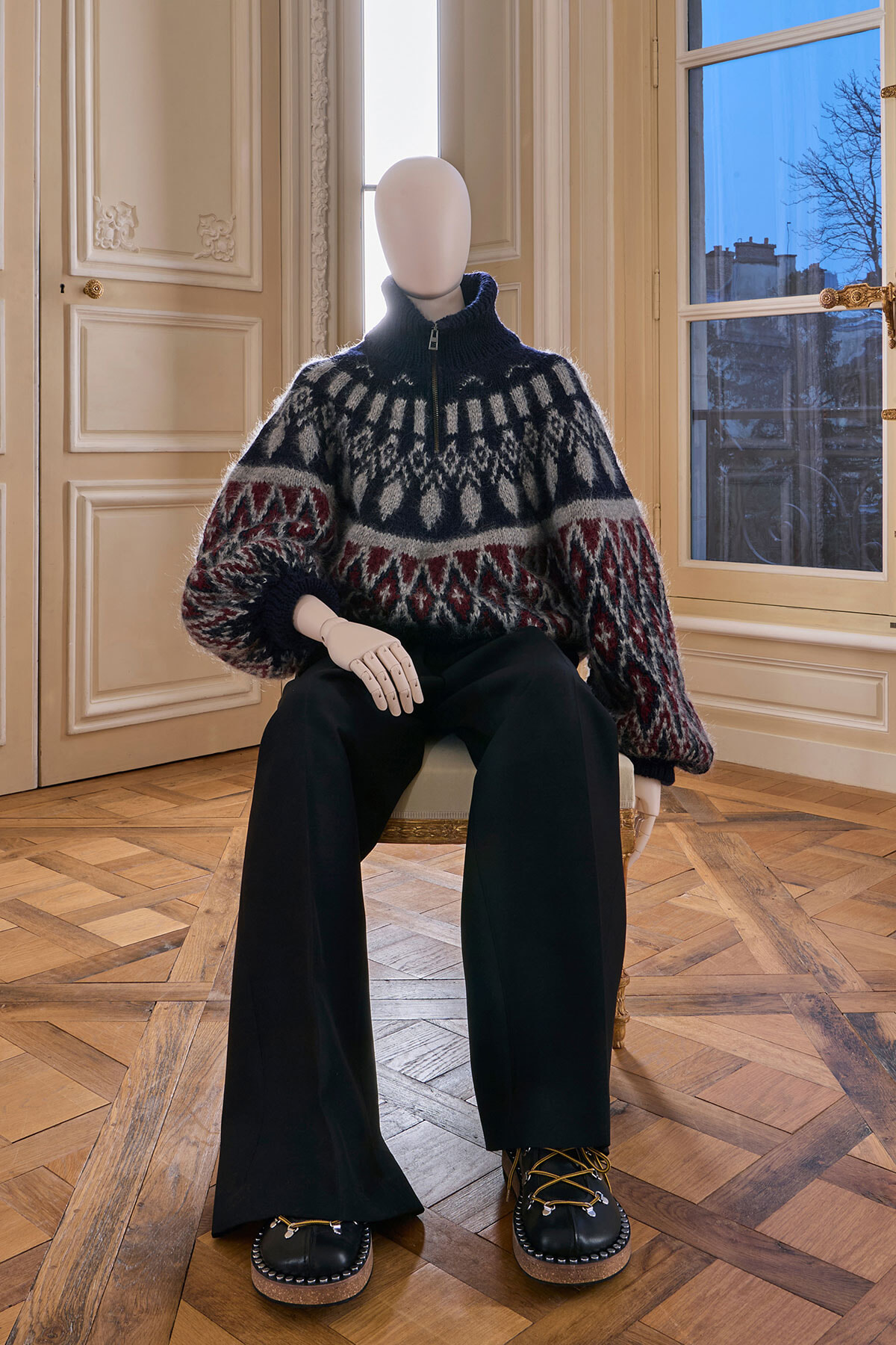 Mannequin seated in an ornate room wearing a chunky navy and gray Nordic-pattern sweater, black wide-leg pants, and dark platform shoes.