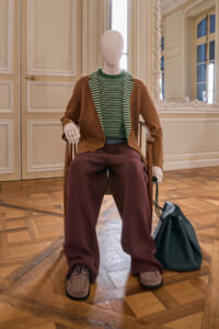 Mannequin seated in a decorative room wearing a brown cardigan over a green striped sweater, burgundy wide-leg pants, plaid loafers, and a teal bag at its side.