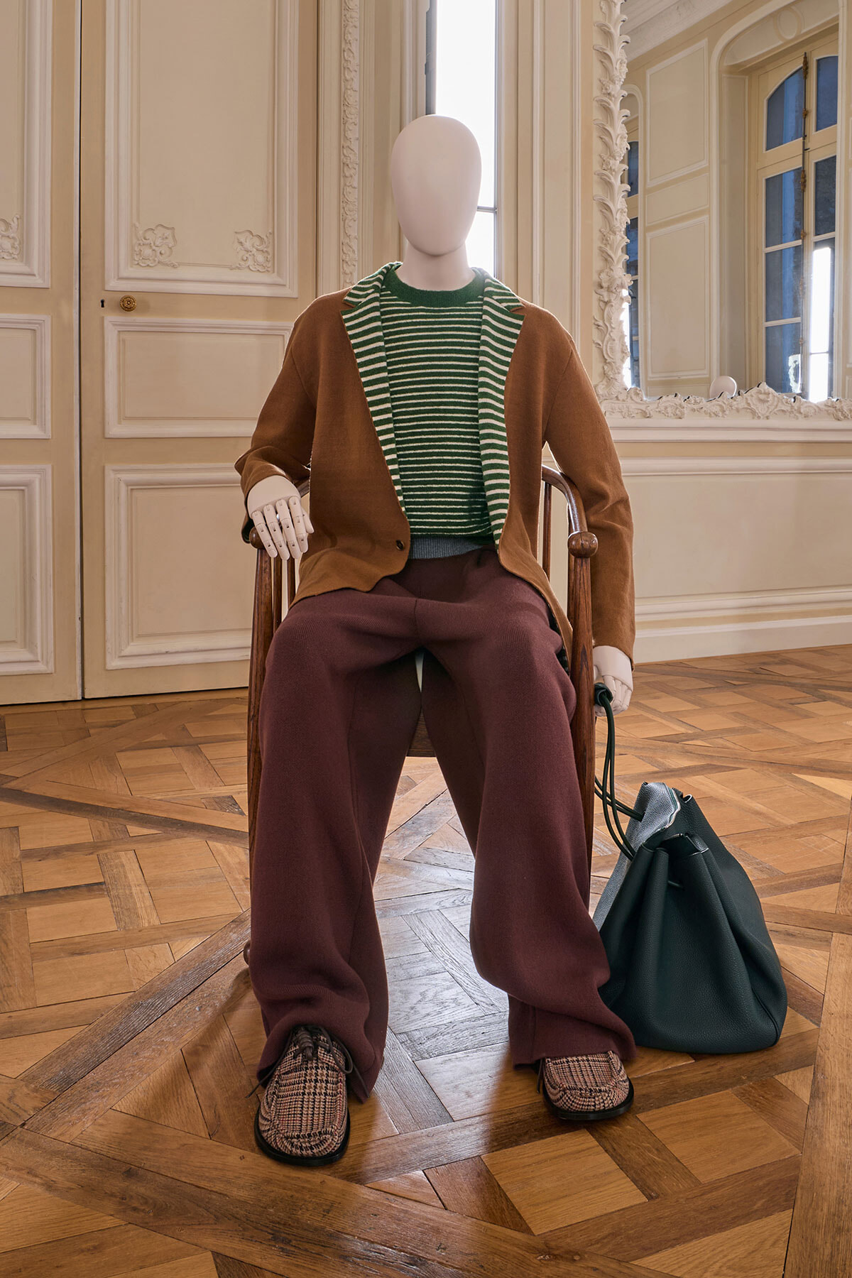 Mannequin seated in a decorative room wearing a brown cardigan over a green striped sweater, burgundy wide-leg pants, plaid loafers, and a teal bag at its side.