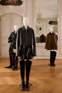 Mannequin wearing a black cropped leather jacket and matching skirt with tall boots in a rococo-style showroom with mirrors and chandelier.
