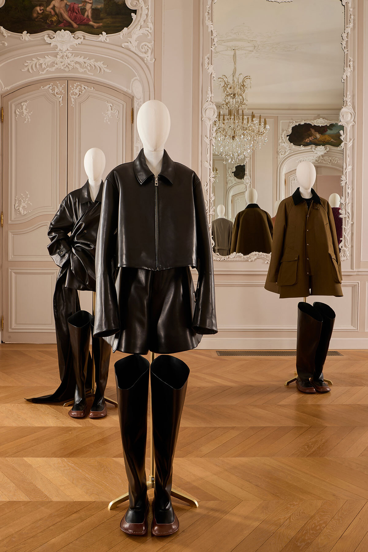 Mannequin wearing a black cropped leather jacket and matching skirt with tall boots in a rococo-style showroom with mirrors and chandelier.