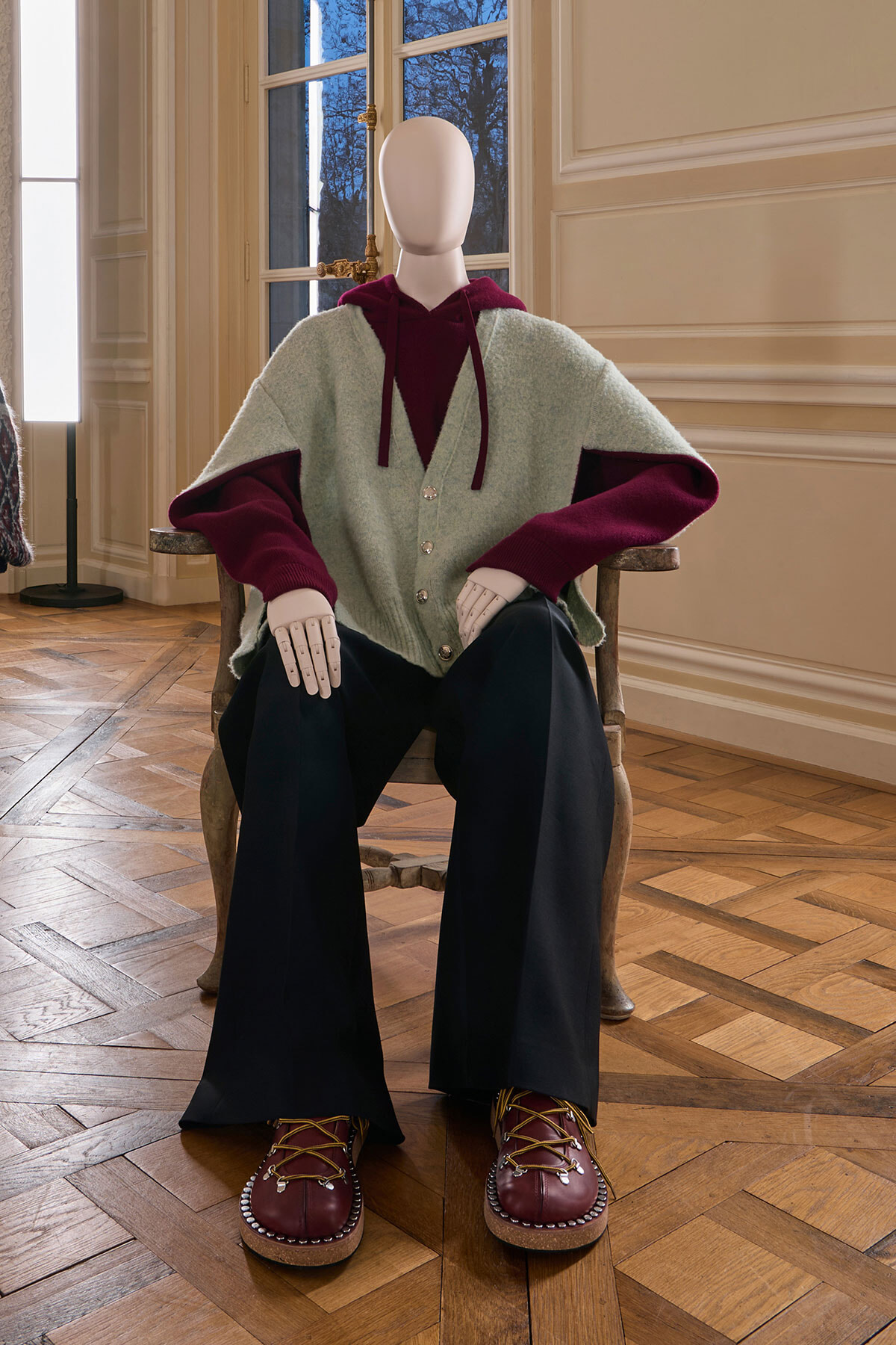 Mannequin seated in a historic room wearing a mint cardigan over burgundy hoodie, dark wide-leg pants, and burgundy lace-up shoes.
