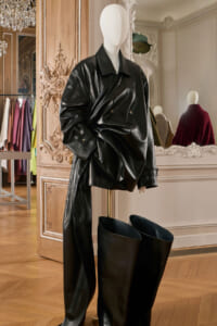 Mannequin wearing an oversized black leather coat and tall black boots in a luxury boutique with ornate wood paneling and mirrors nearby.