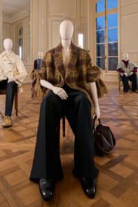 Mannequin in a brown plaid oversized coat with wide-leg black pants and black shoes, seated with a brown handbag in an ornate gallery setting.