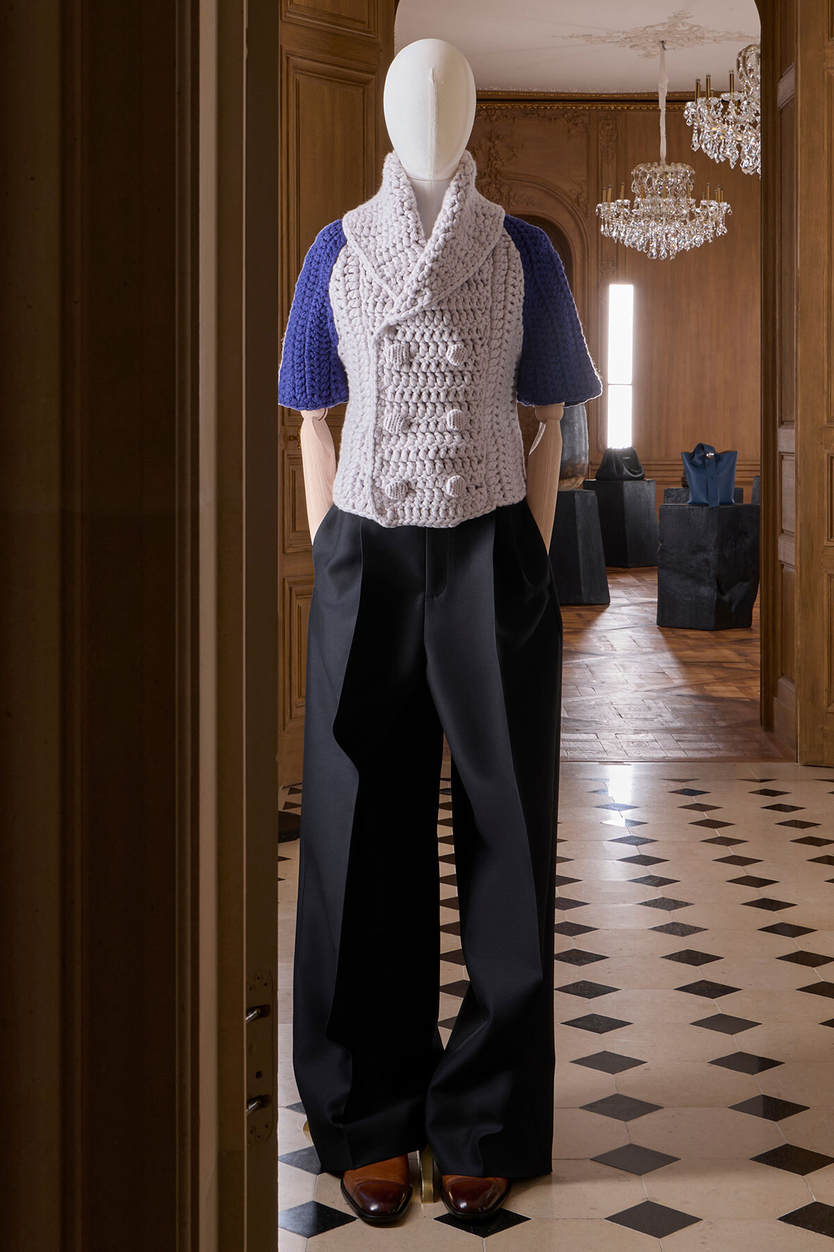 Mannequin wearing a chunky knit beige vest over blue sleeves and black wide-leg pants in an elegant showroom setting.