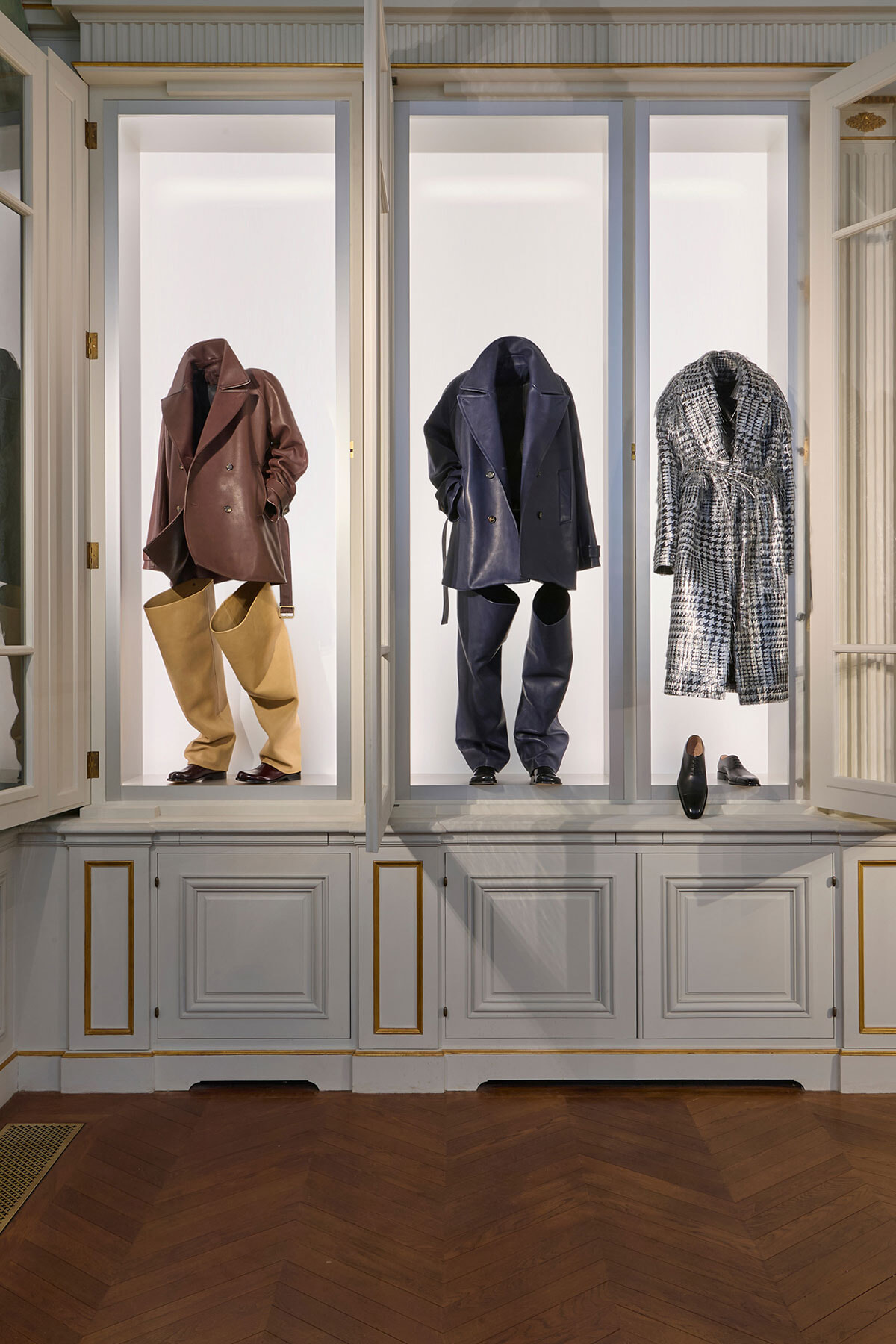 Three mannequins in glass display windows showcasing outfits: brown coat with tan wide pants, navy coat with matching pants, and a black-and-white houndstooth belted coat with shoes nearby.