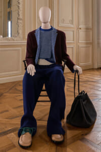 Mannequin seated in an ornate room wearing a burgundy blazer, blue striped sweater, navy wide-leg pants, tan loafers, with a black bag beside it.