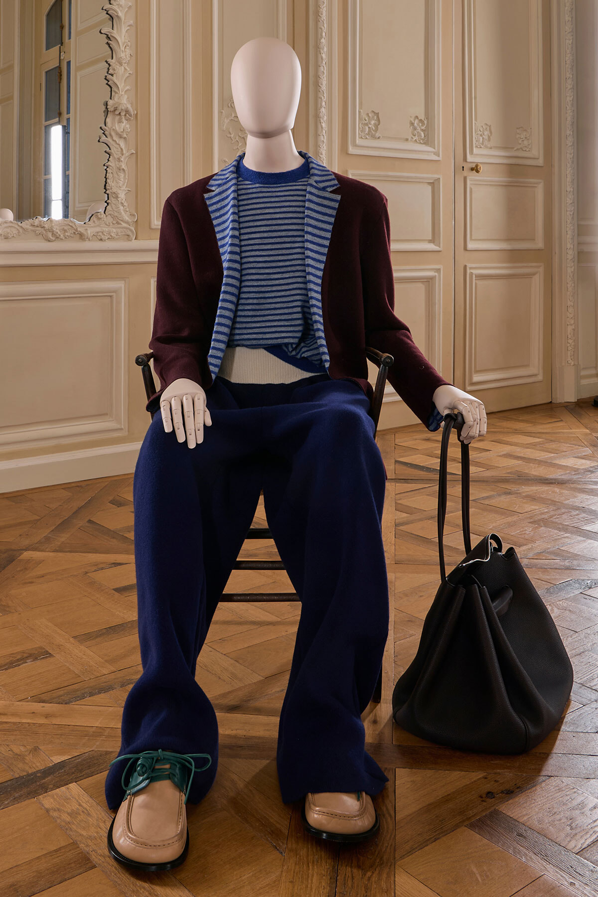 Mannequin seated in an ornate room wearing a burgundy blazer, blue striped sweater, navy wide-leg pants, tan loafers, with a black bag beside it.
