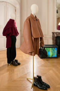 Mannequin in a tan oversized leather coat and skirt, carrying a black handbag; black boots on the wooden floor in an elegant showroom.