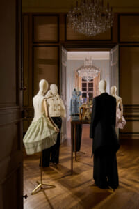 Mannequins in a luxury boutique display with elegant outfits, crystal chandeliers, and a mirrored room beyond the doorway.