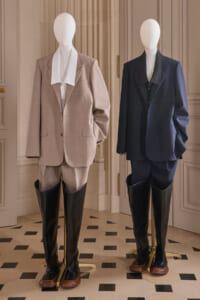 Two mannequins in a showroom wearing tailored suits with tall black riding boots, beige on the left and navy on the right.