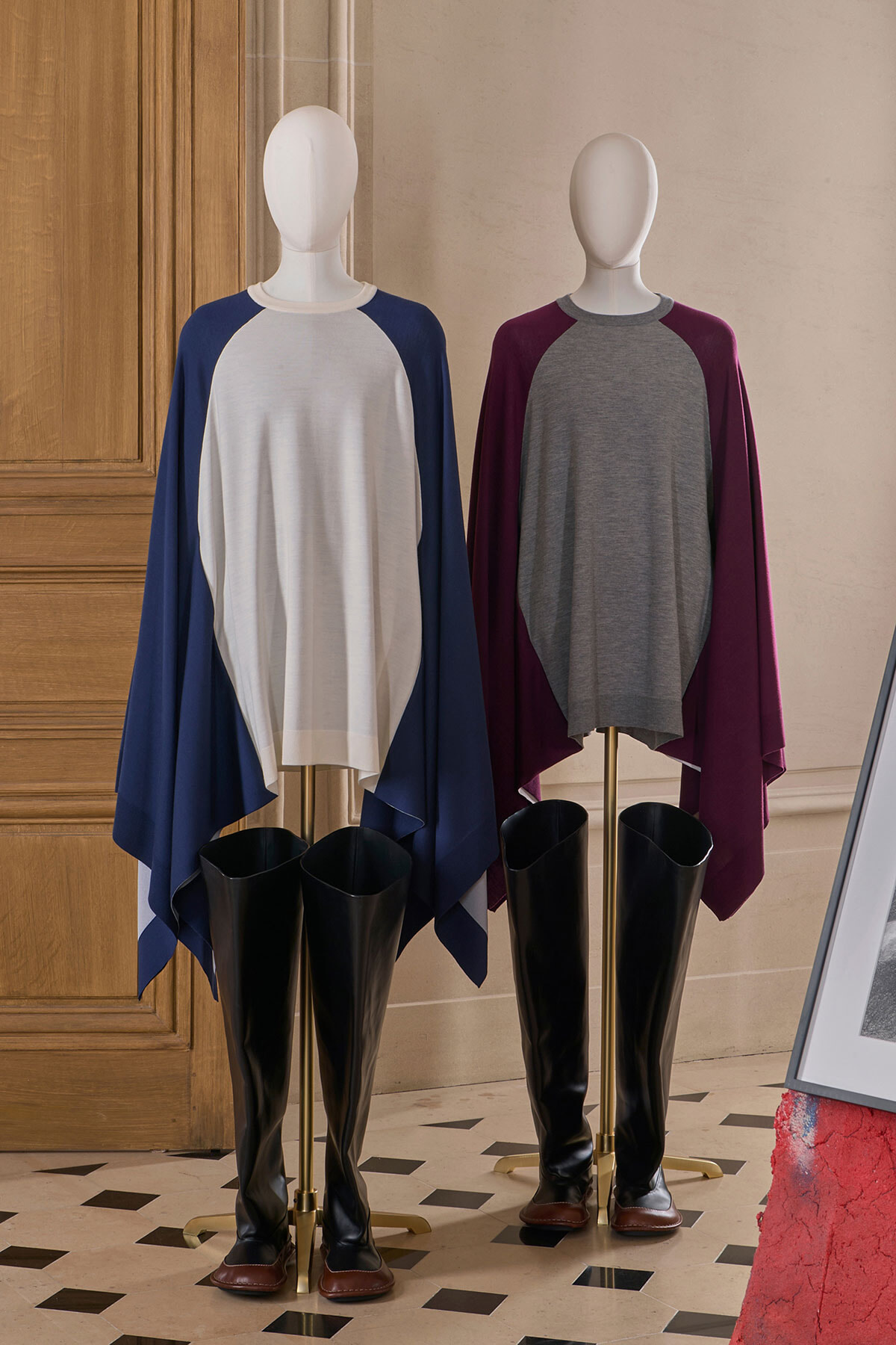 Two mannequins wearing color‑block ponchos and knee‑high black boots, on display in a boutique with wooden panels behind them.