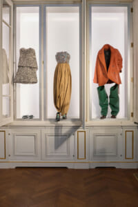 Three outfits displayed in glass-front cabinets: a textured strapless dress on the left, a beaded bodice with gold draped skirt in the center, and an orange coat with green pants on the right.