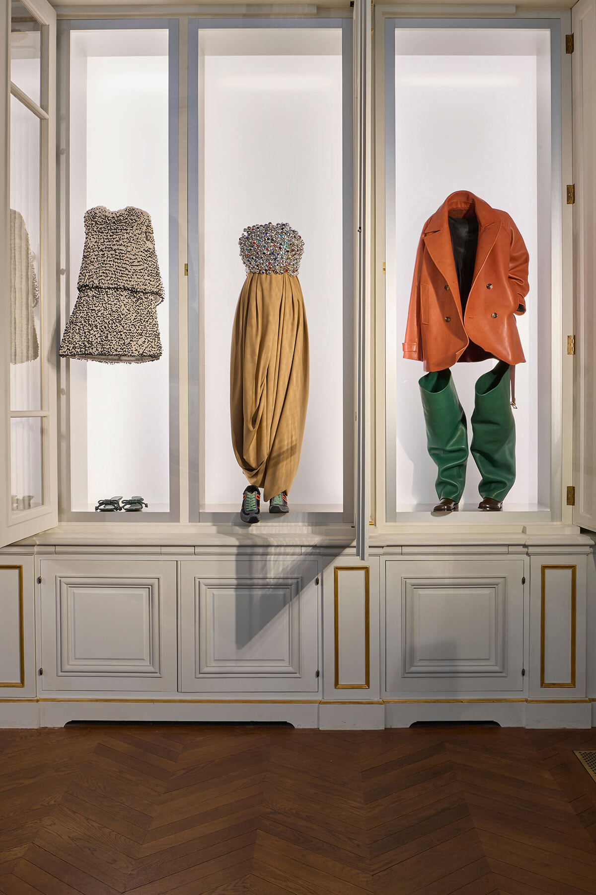 Three outfits displayed in glass-front cabinets: a textured strapless dress on the left, a beaded bodice with gold draped skirt in the center, and an orange coat with green pants on the right.