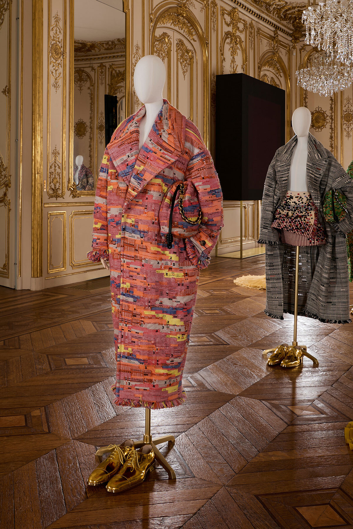 Mannequin wearing a pink, multicolored textured coat with matching fringe, in an ornate gold-trimmed room with wooden parquet flooring and gold shoes beneath it.