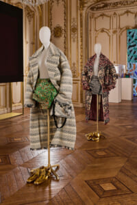 Two mannequins wearing oversized patterned coats and metallic gold shoes in a gilded, ornate room display.