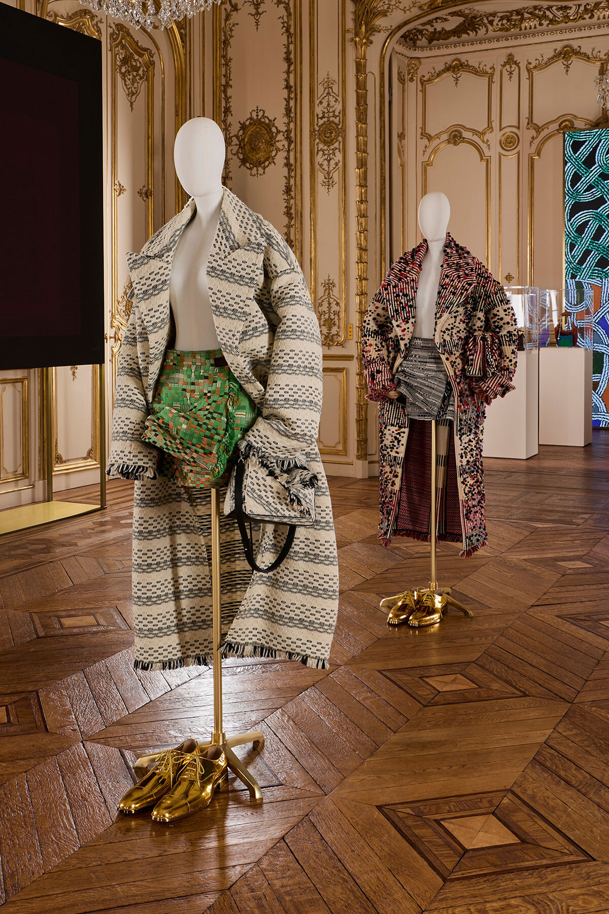 Two mannequins wearing oversized patterned coats and metallic gold shoes in a gilded, ornate room display.