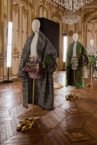 Mannequins display stylish outerwear in an ornate gilded room; front mannequin wears a grey plaid coat with a multicolored knit bag nearby.