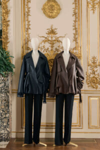 Two mannequins wearing oversized leather jackets (black and brown) on display in an ornate gilded room, black trousers, luxurious decor backdrop.